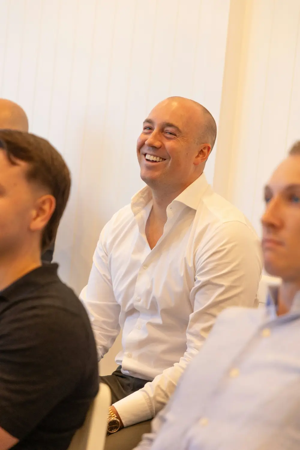 A man in a white open-collar shirt laughs while seated among other people in a room.