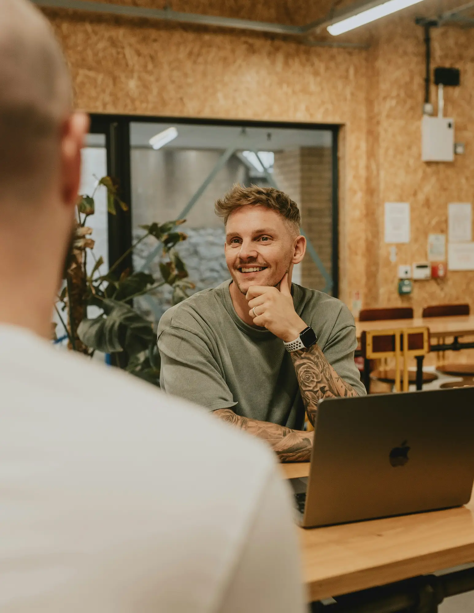 A man smiles and listens attentively during a one-on-one conversation across a table, with a laptop open in front of him.