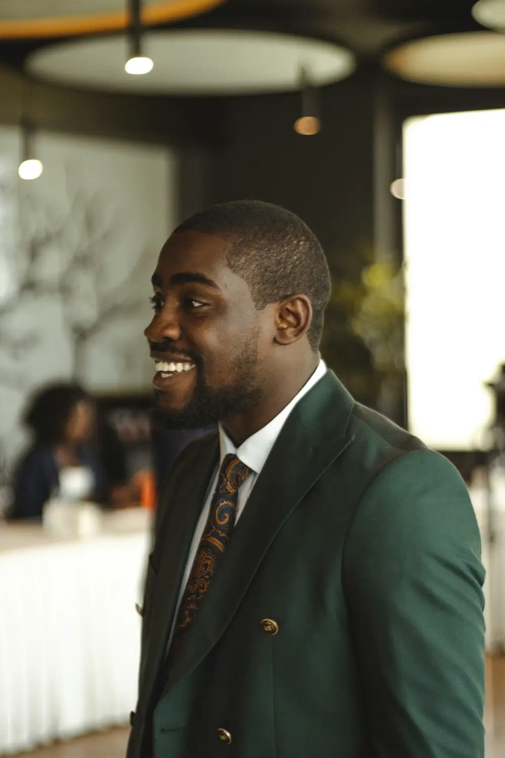 A smiling man in a green double-breasted blazer and patterned tie looks off to the side.
