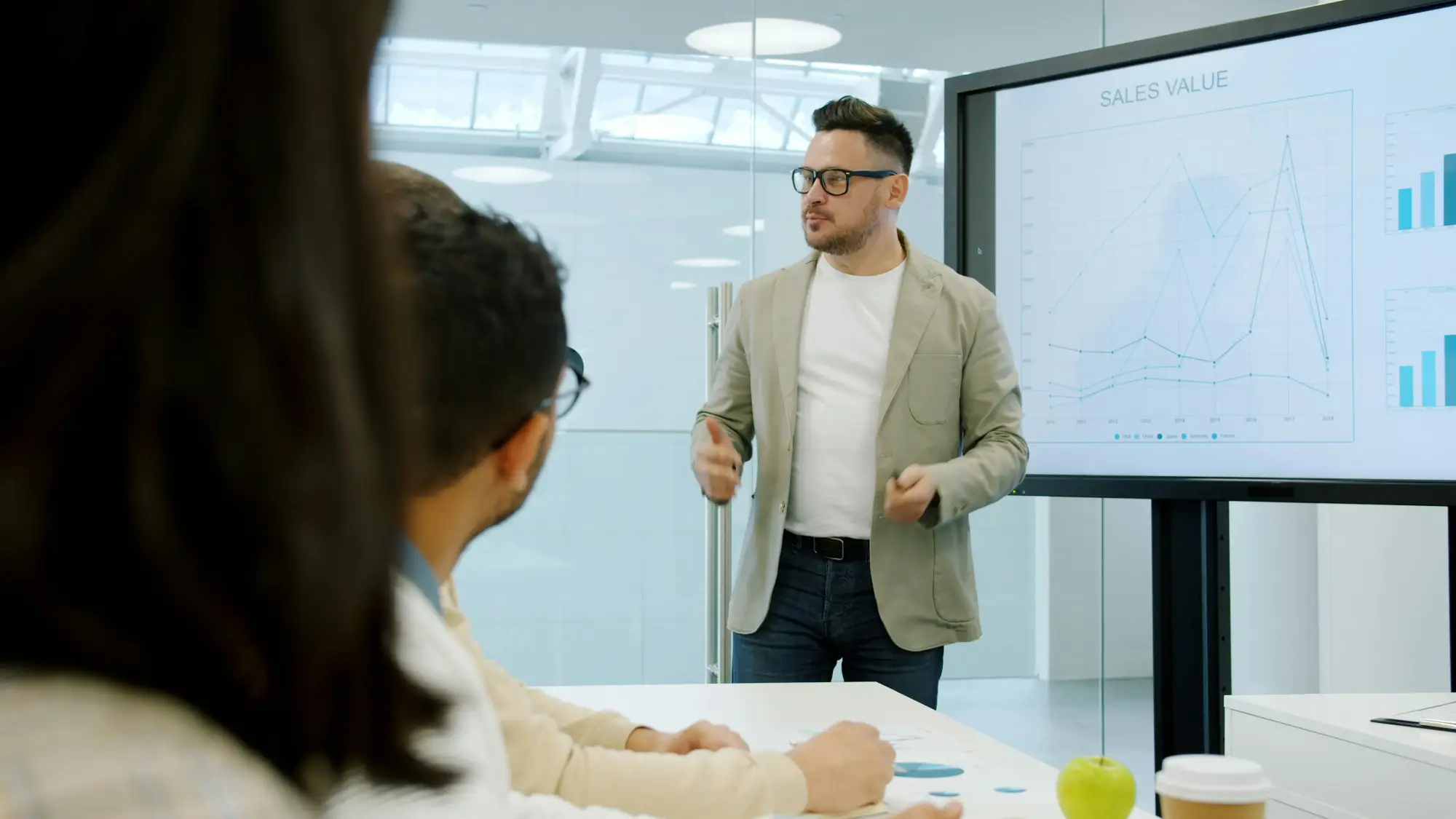 A sales leader presents a performance chart on a wall-mounted screen to colleagues seated around a conference table.