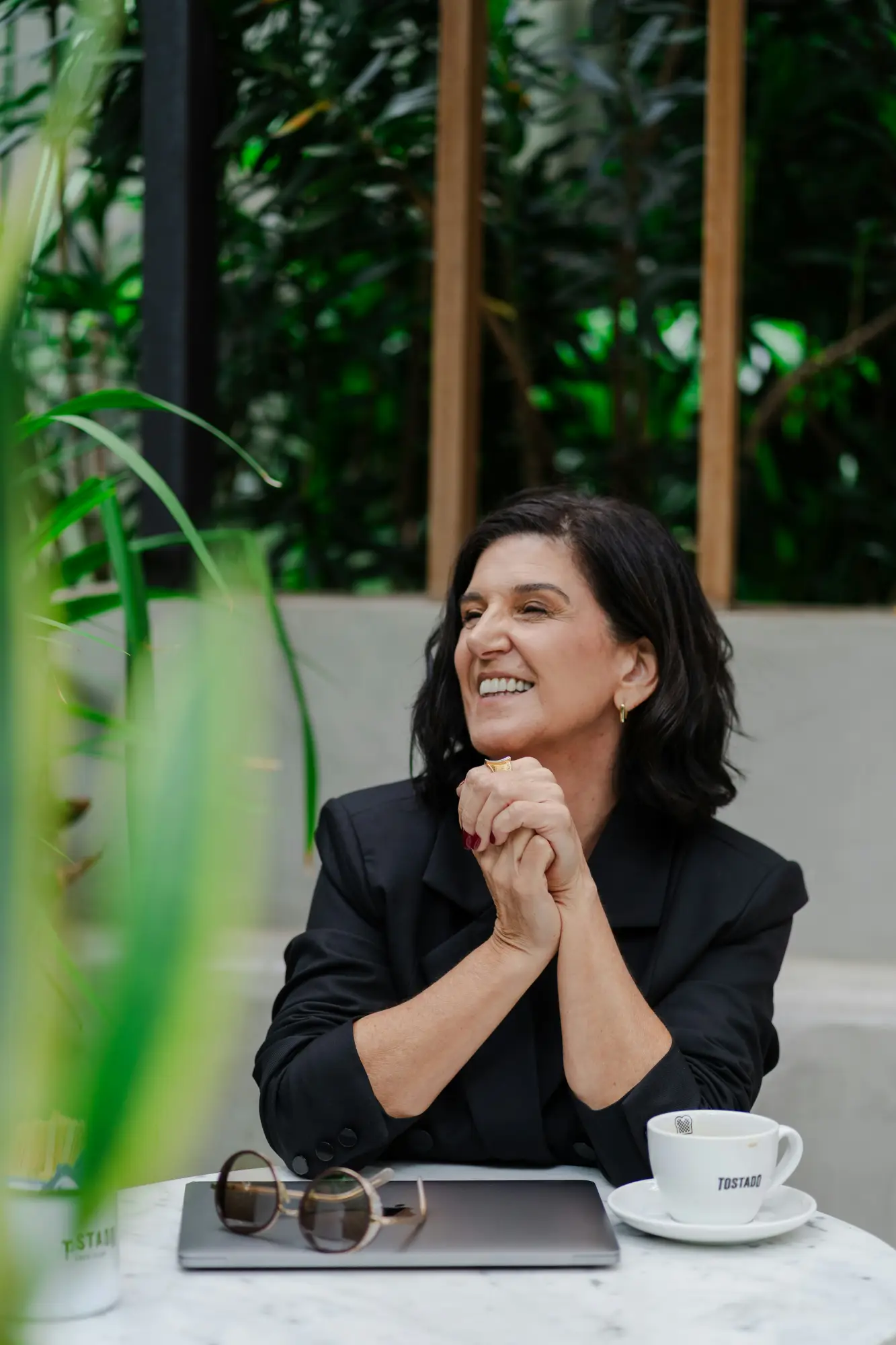 A woman in a black blazer smiles with her hands clasped at a table with a laptop, sunglasses and a coffee cup.