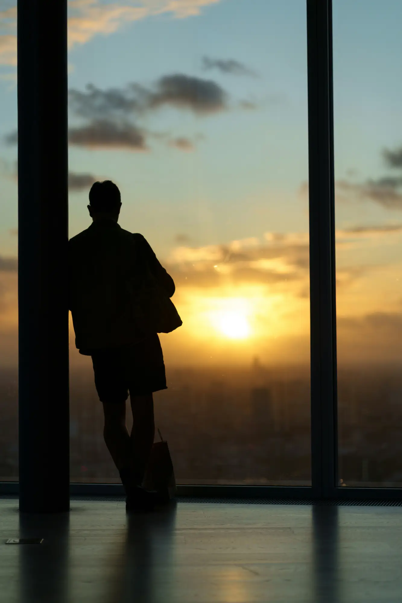 Silhouette of a person leaning against a column and looking out a floor-to-ceiling window at a city skyline at sunset.