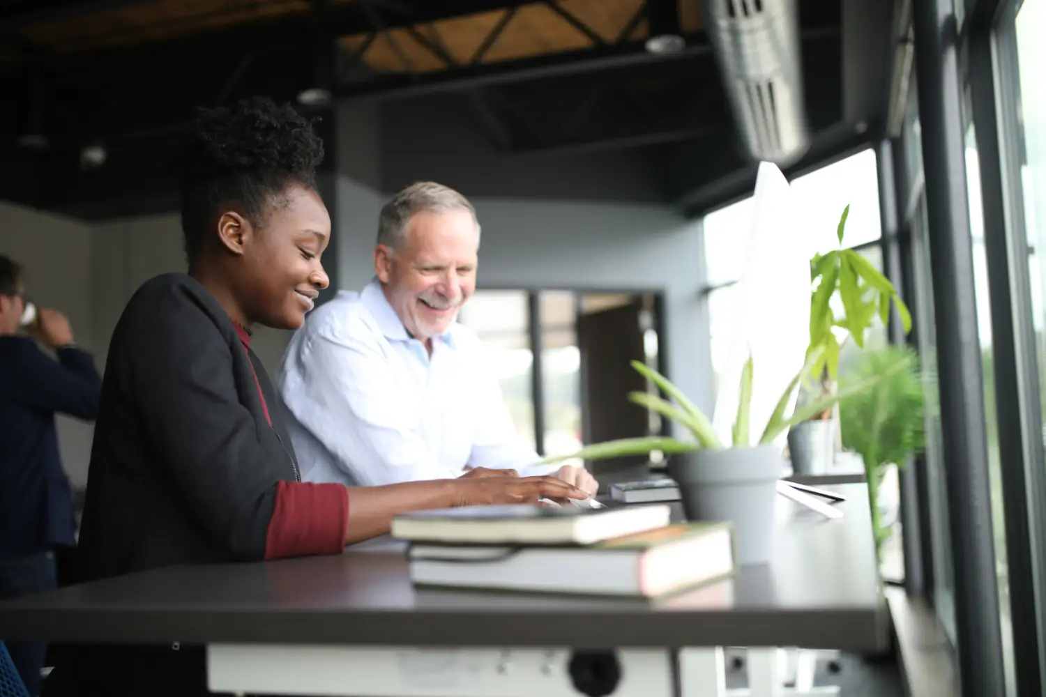 Two colleagues work side by side at a desk, smiling as one types and the other looks on.