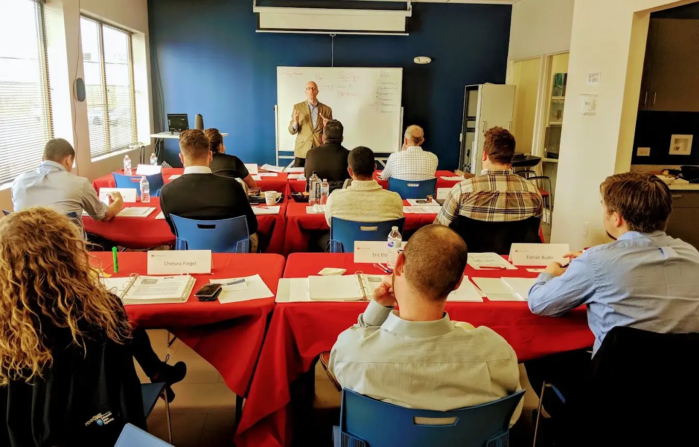Sales Evolution instructor leads a sales training session at a whiteboard while participants follow along at tables with binders and notes.